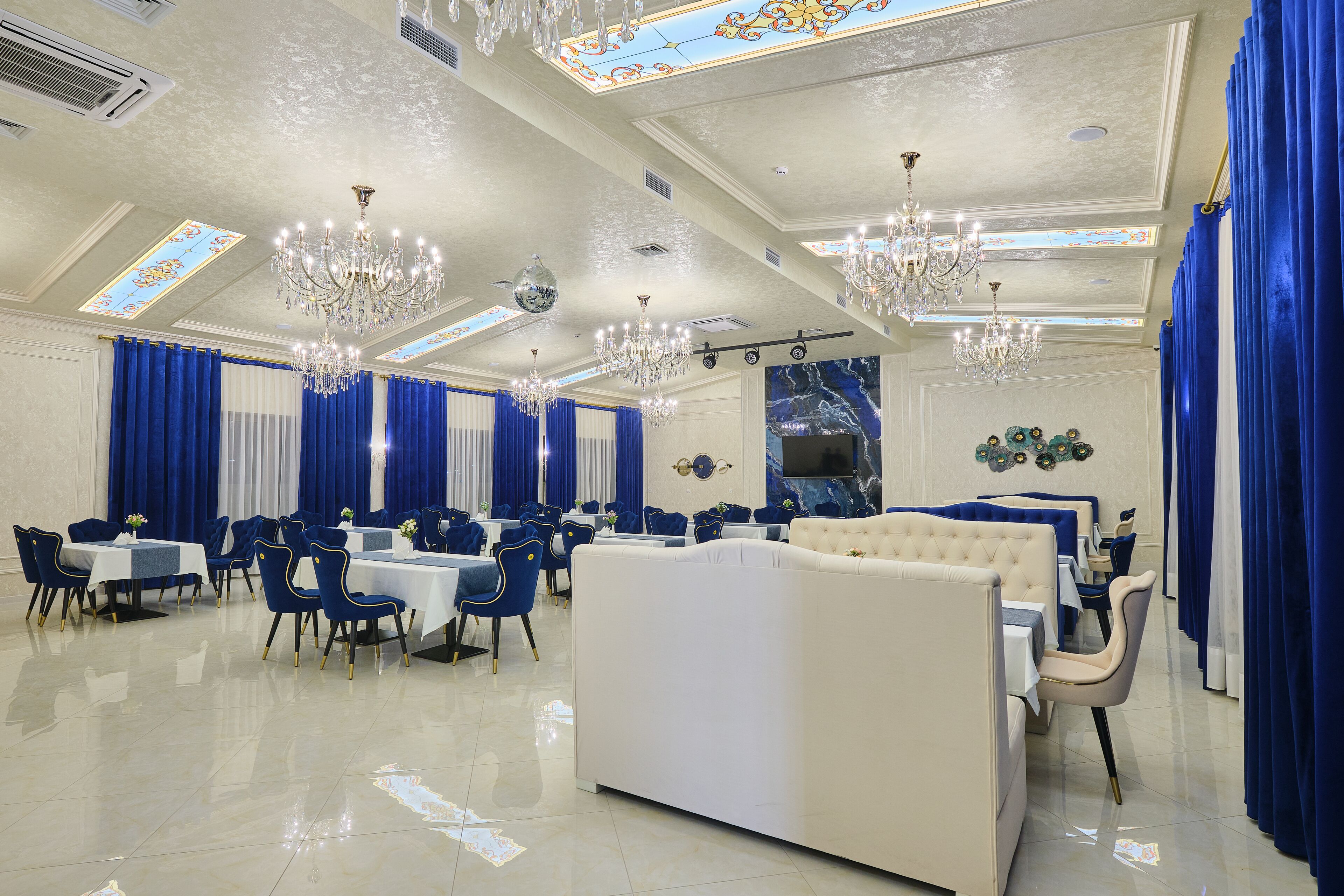 A spacious dining area with high ceilings, chandeliers, and blue curtains. Tables are set with white tablecloths and blue chairs.