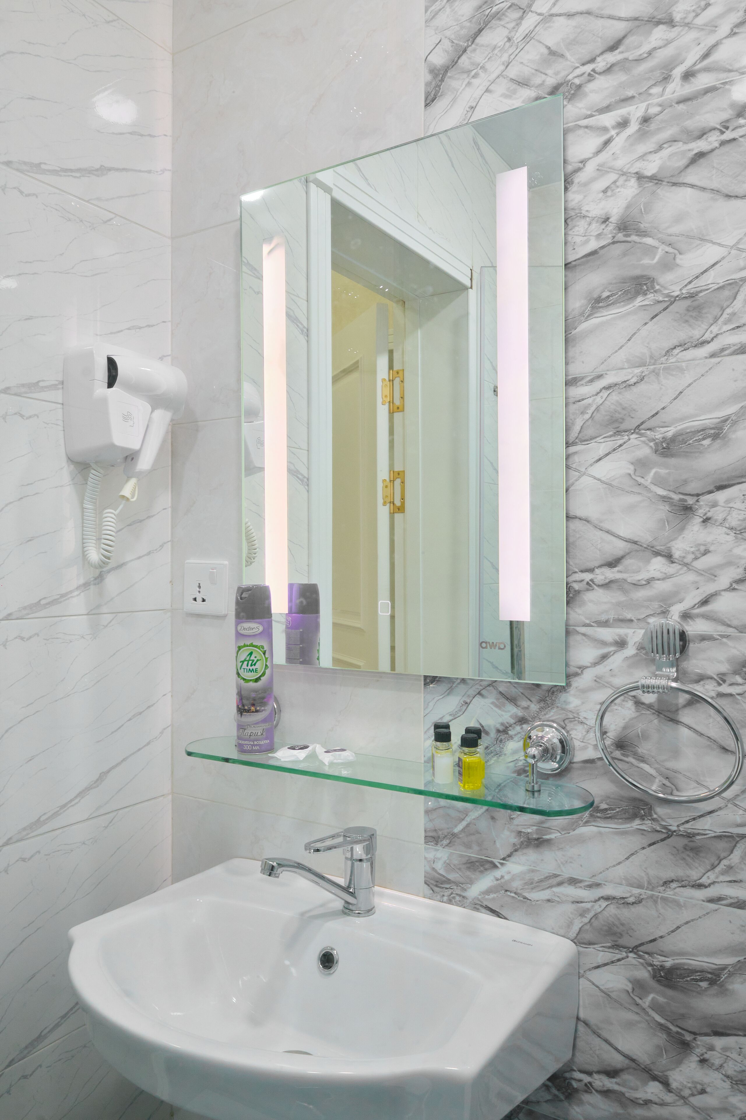 A bathroom with a white sink, a glass shelf with toiletries, a hairdryer, and a marble-patterned wall.