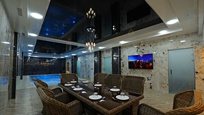 A modern dining area with a long table set for six, surrounded by wicker chairs, a chandelier, and a large TV screen. - Samaria Hotel and Spa (Samarkand)