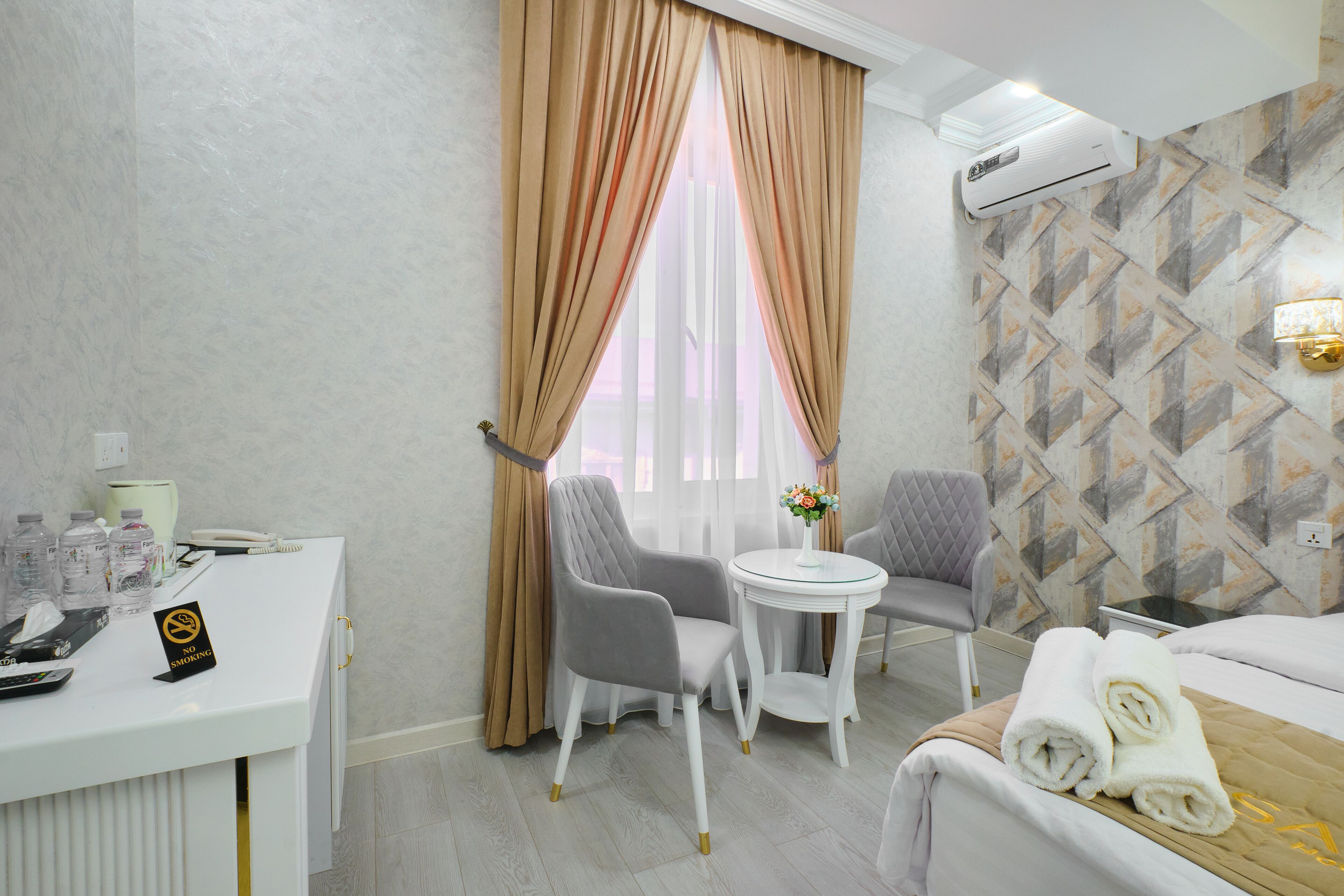 Quarto de hotel com escrivaninha, duas cadeiras, uma mesinha redonda com um vaso e uma cama com toalhas, além de um papel de parede estampado.