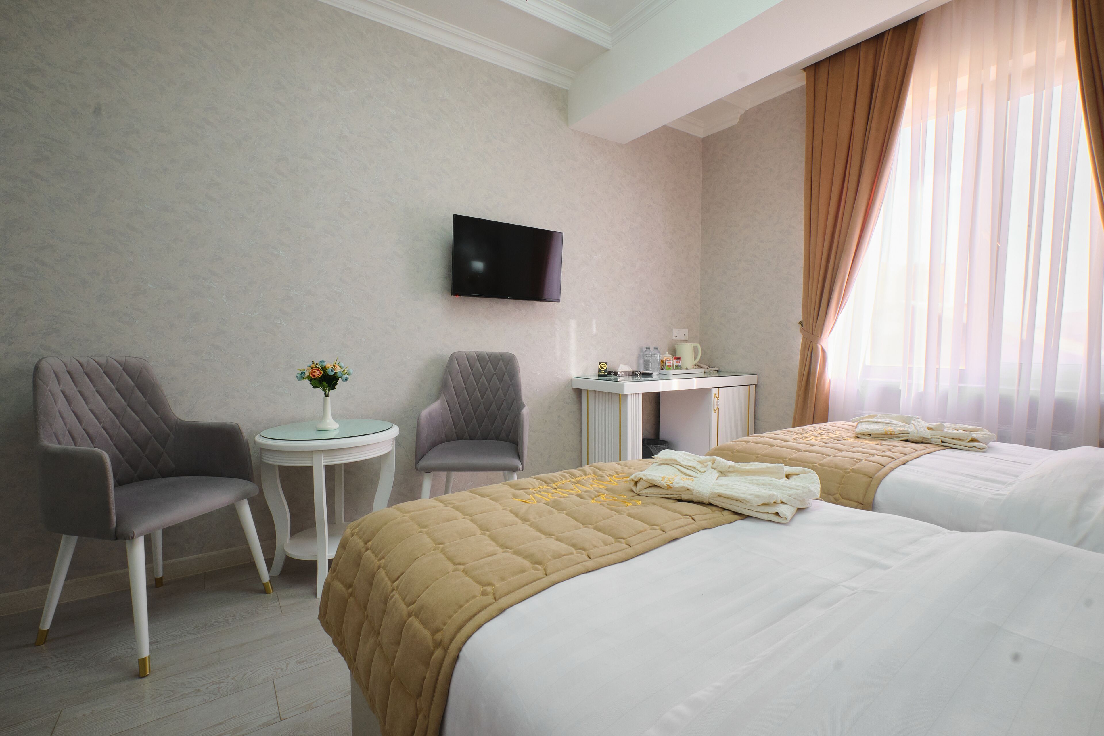 A hotel room with two beds, a TV mounted on the wall, a small round table with a vase of flowers, and a desk with a chair.