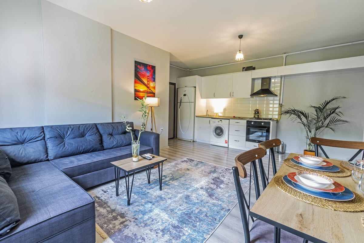 A modern living room with a sofa and a dining table set for two, a kitchen in the background, and a painting on the wall.