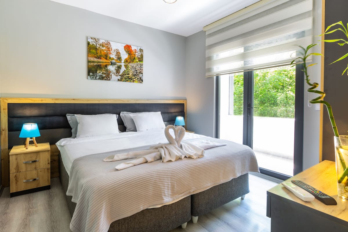 A modern bedroom with a large bed, a nightstand with a lamp, a painting on the wall, and a view of greenery through the window.