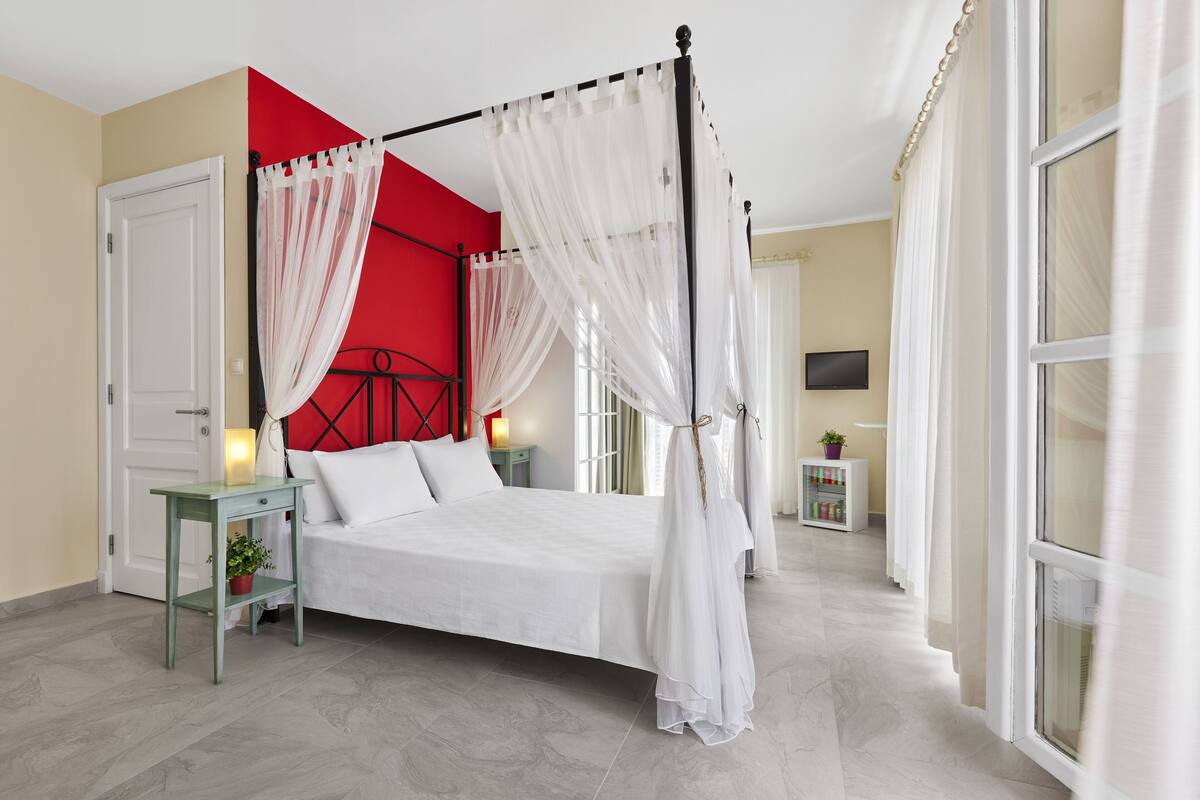 A four-poster bed with white bedding, a red accent wall, and a small bedside table with a lamp.