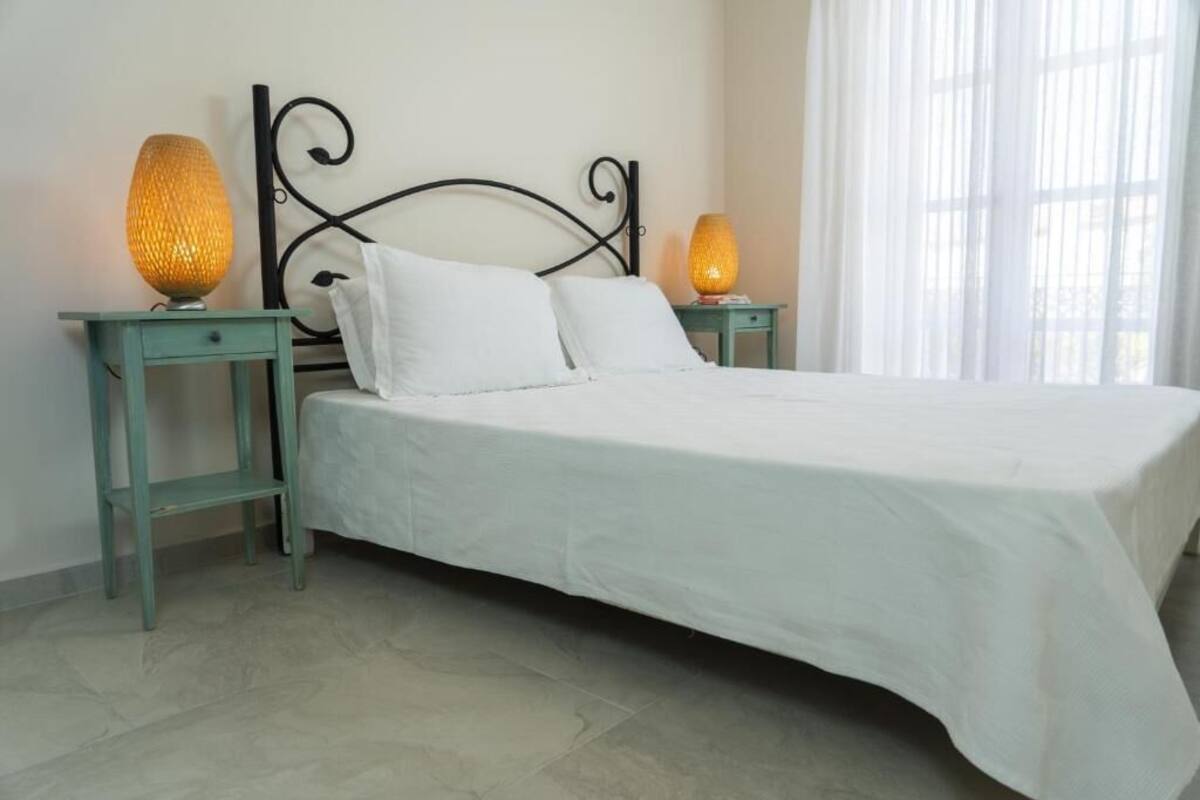 A neatly made bed with white linens, two bedside tables with lamps, and a window with sheer curtains.
