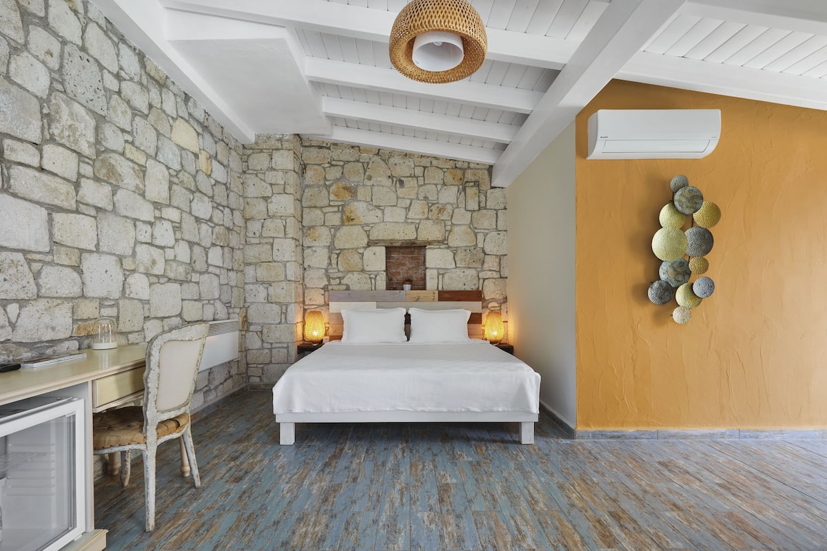 A bedroom with a stone wall, a bed with white bedding, a desk with a chair, and a wall-mounted air conditioner.