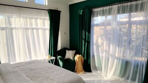 Ein Hotelzimmer mit einem Bett, einem grünen Sessel, einem kleinen Tisch mit einer Kerze und durchsichtigen Vorhängen an den Fenstern.
