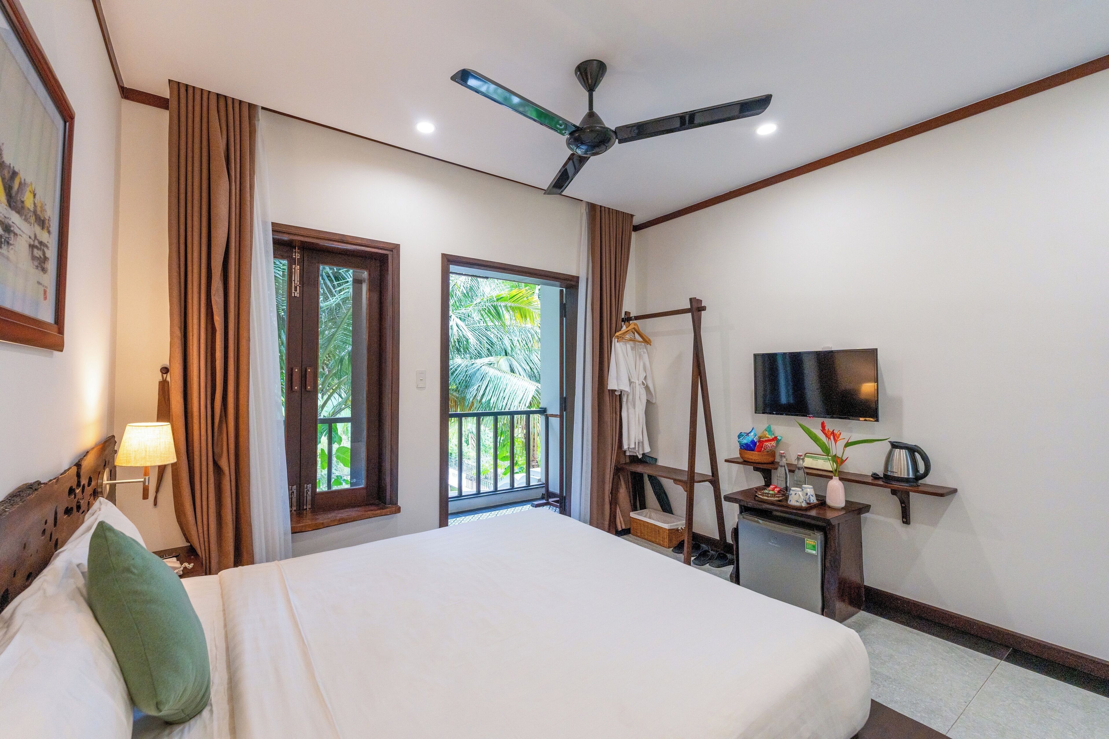 Une chambre d’hôtel comprenant un lit, un ventilateur de plafond, une télévision, un bureau avec une machine à café et un balcon avec des rideaux.