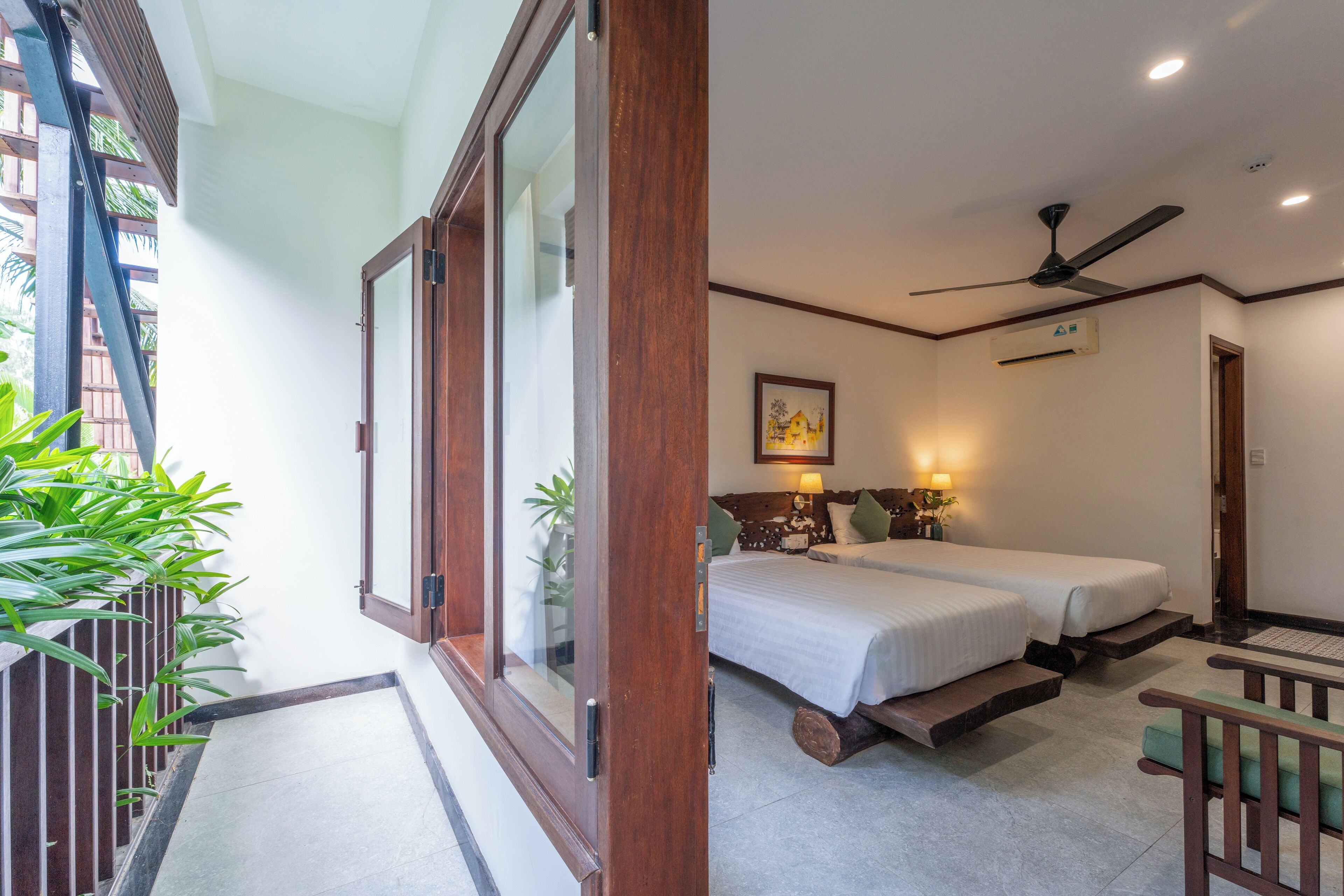 Habitación de hotel con balcón, dos camas, ventilador de techo y una pequeña zona de estar.