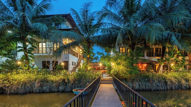 A wooden bridge leading to a building with a tiled roof, surrounded by palm trees and a calm water body.