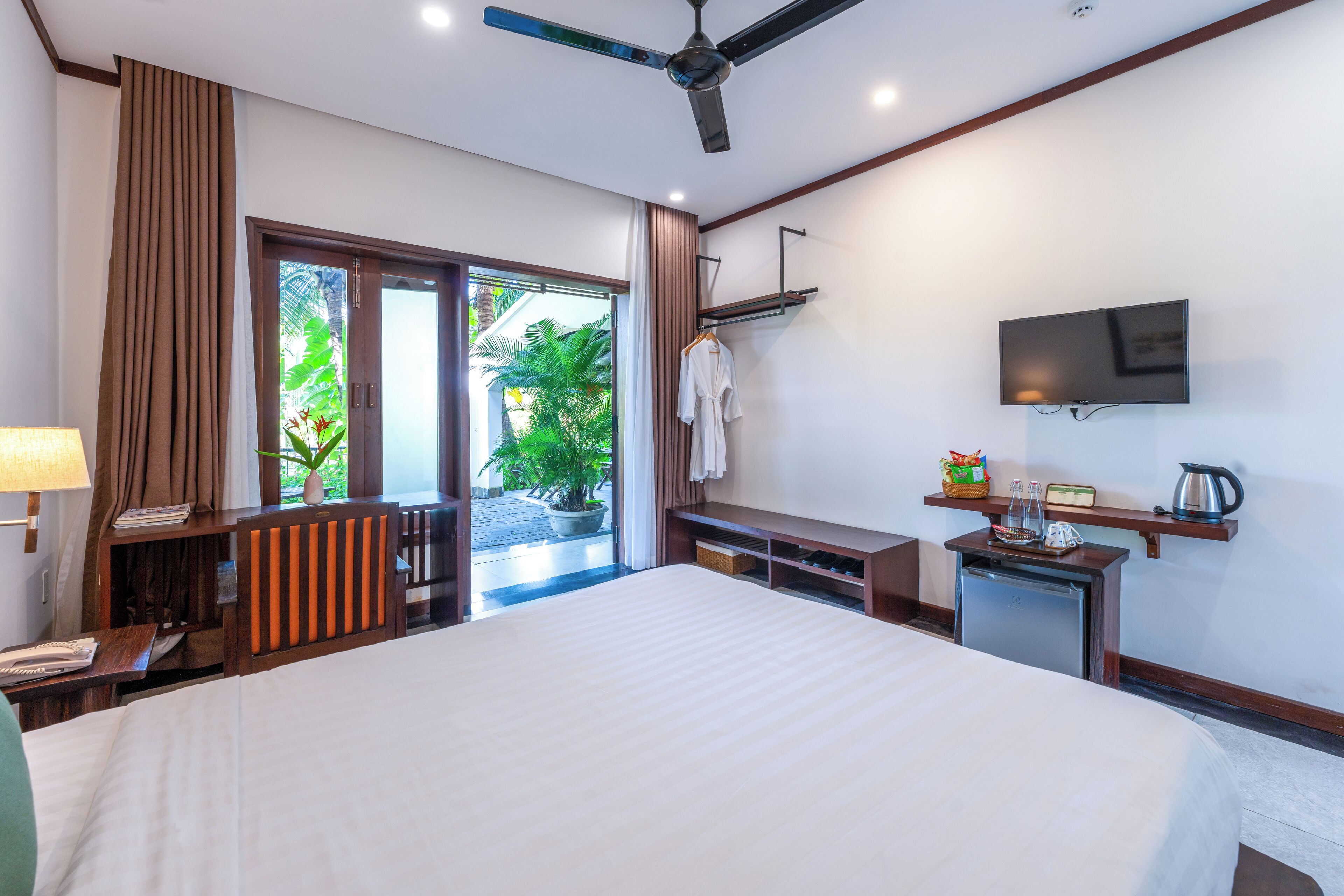 A hotel room with a large bed, a ceiling fan, a TV, a small kitchenette, and a view of greenery outside.