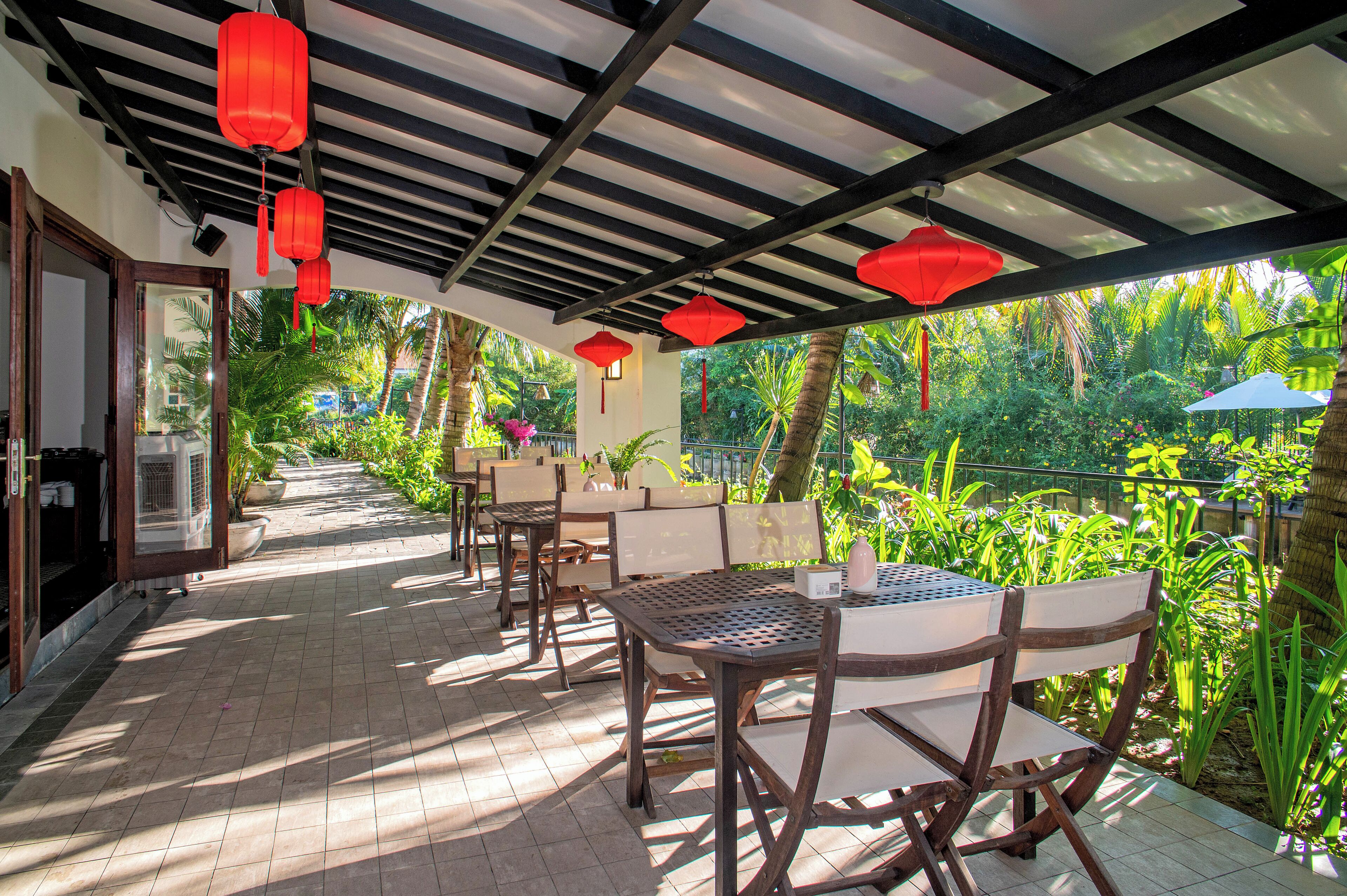 A covered outdoor seating area with tables and chairs, red lanterns hanging from the ceiling, and a view of lush greenery.