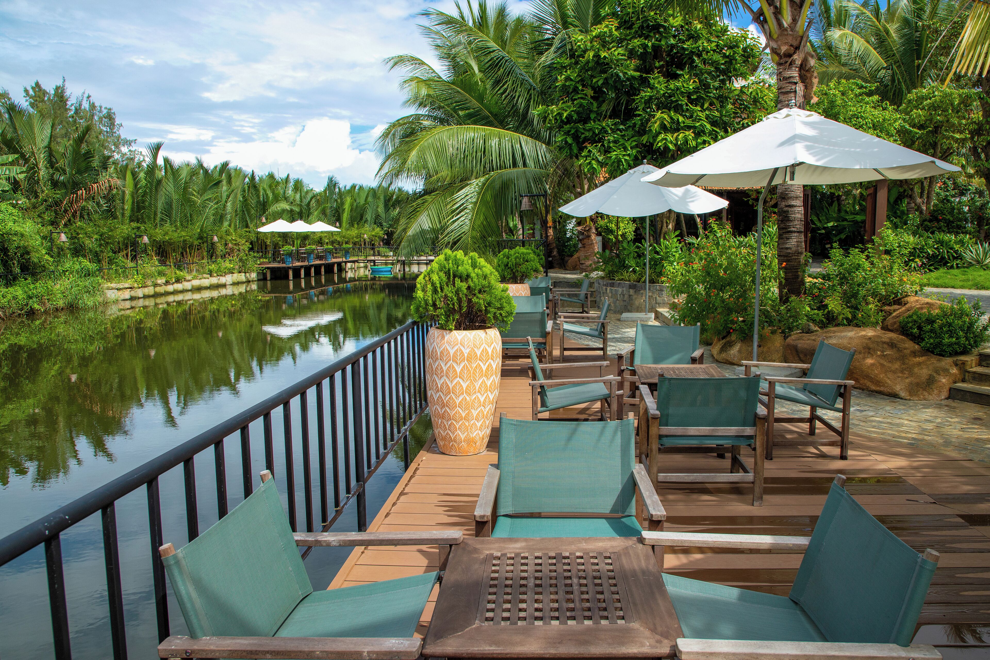 Une terrasse en bois, agrémentée de chaises vertes à coussins et de parasols blancs, qui offre une vue sur une étendue d’eau paisible et une végétation luxuriante.
