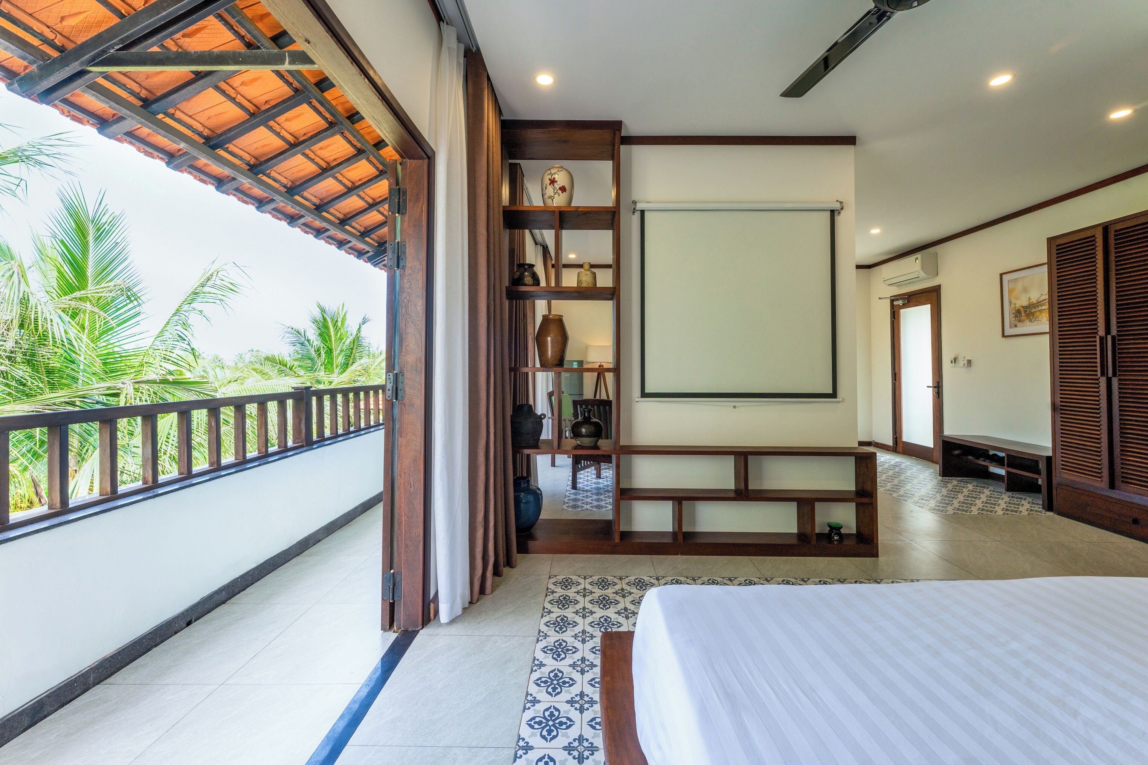 Habitación de hotel con balcón, una cama, repisas de madera con adornos y vista a palmeras.