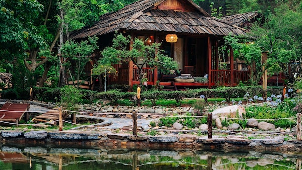 Uma casa rústica de madeira com telhado de palha, rodeada por vegetação exuberante e um lago que reflete a construção.