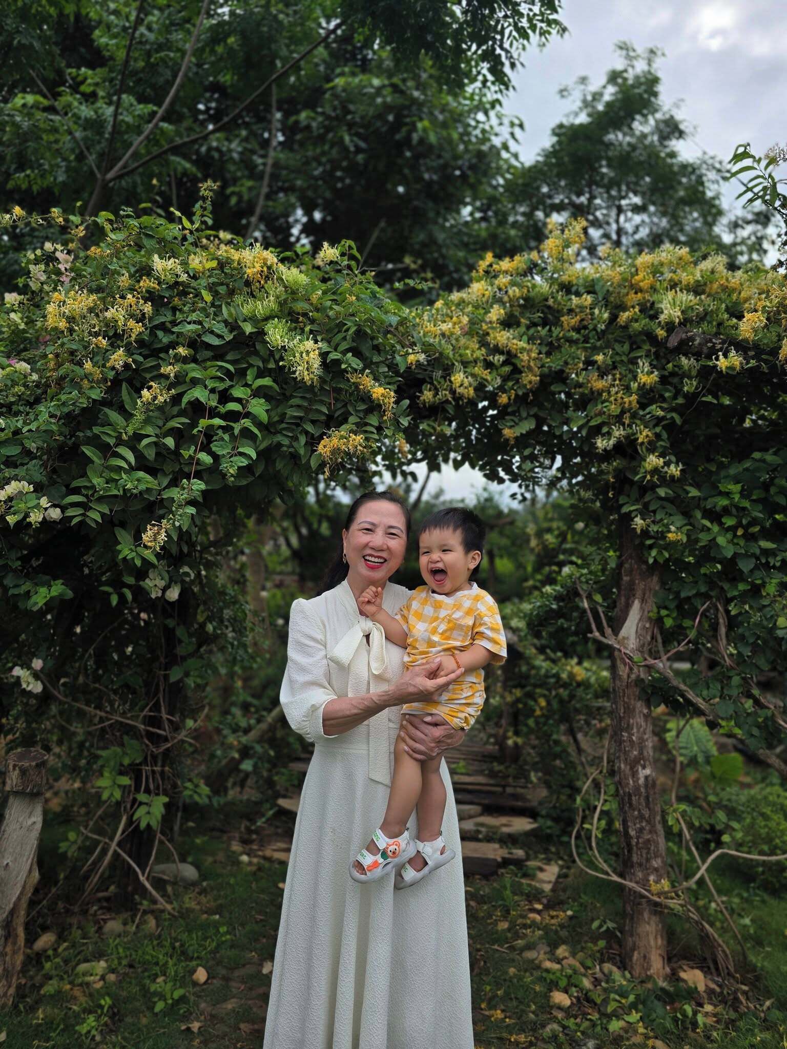 A person holding a child under a leafy archway in a natural setting.