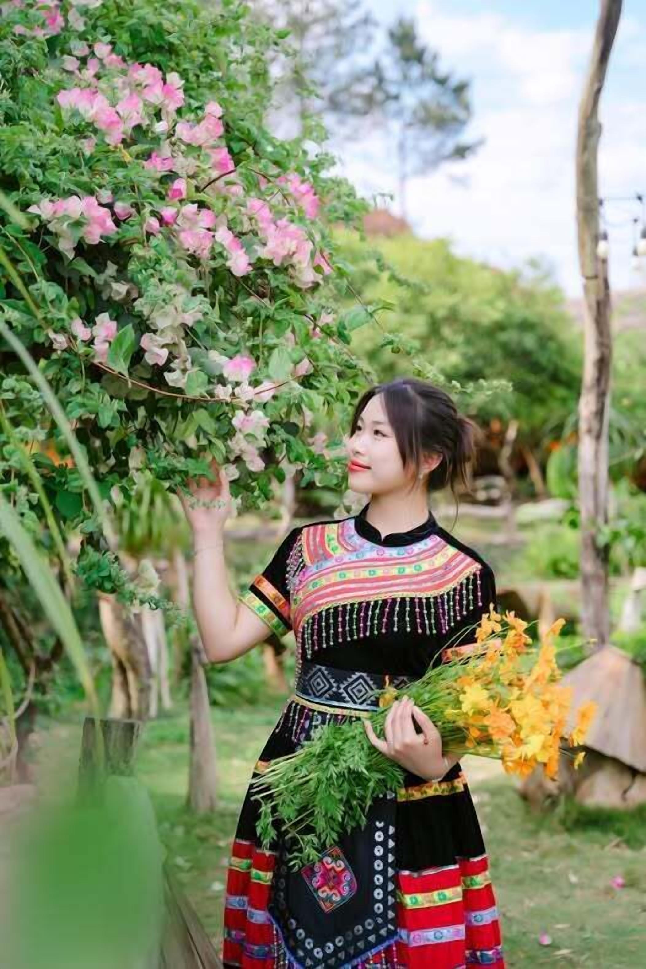 Una persona vestida con traje tradicional sostiene flores y un ramo de follaje, de pie en un jardín con árboles y una cerca de madera.