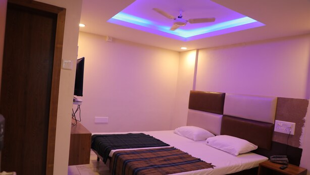A bedroom with a ceiling fan, a bed with two pillows, a bedside table with a remote control, and a wooden nightstand. - kapila residency (Bengaluru)