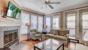 A spacious living room with a fireplace, a ceiling fan, a large TV, and a balcony with a railing. - Steps to Two Oceanfront Pools | Walk to Dining | Grand Pavilion (Isle of Palms)