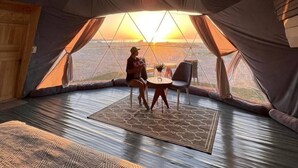 Una tienda de campaña con una mesa central y sillas, un candelabro y vistas al atardecer.