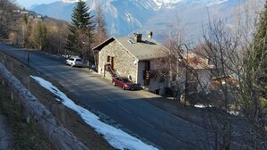 Ein Steinhaus mit geneigtem Dach, ein Auto davor und eine Straße, die zu einem Gebirgszug führt.