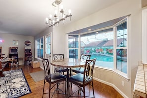 A dining area with a round table and chairs, a chandelier, arcade games, and a view of a swimming pool. - Fun-filled, modern home with game room, heated pool, hot tub, grill (Chandler)