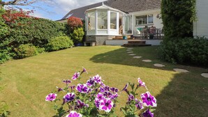 Une maison avec une véranda, une pelouse bien entretenue et un jardin agrémenté de fleurs violettes.