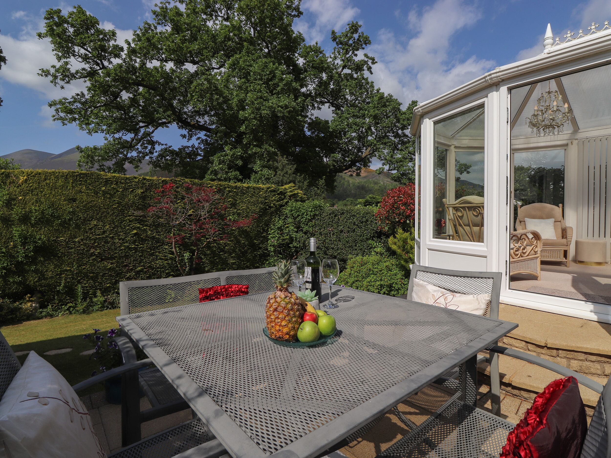 Ein Terrassentisch mit einer Ananas, Äpfeln und Weingläsern, umgeben von Korbstühlen und einem weißen Gebäude mit Balkon.
