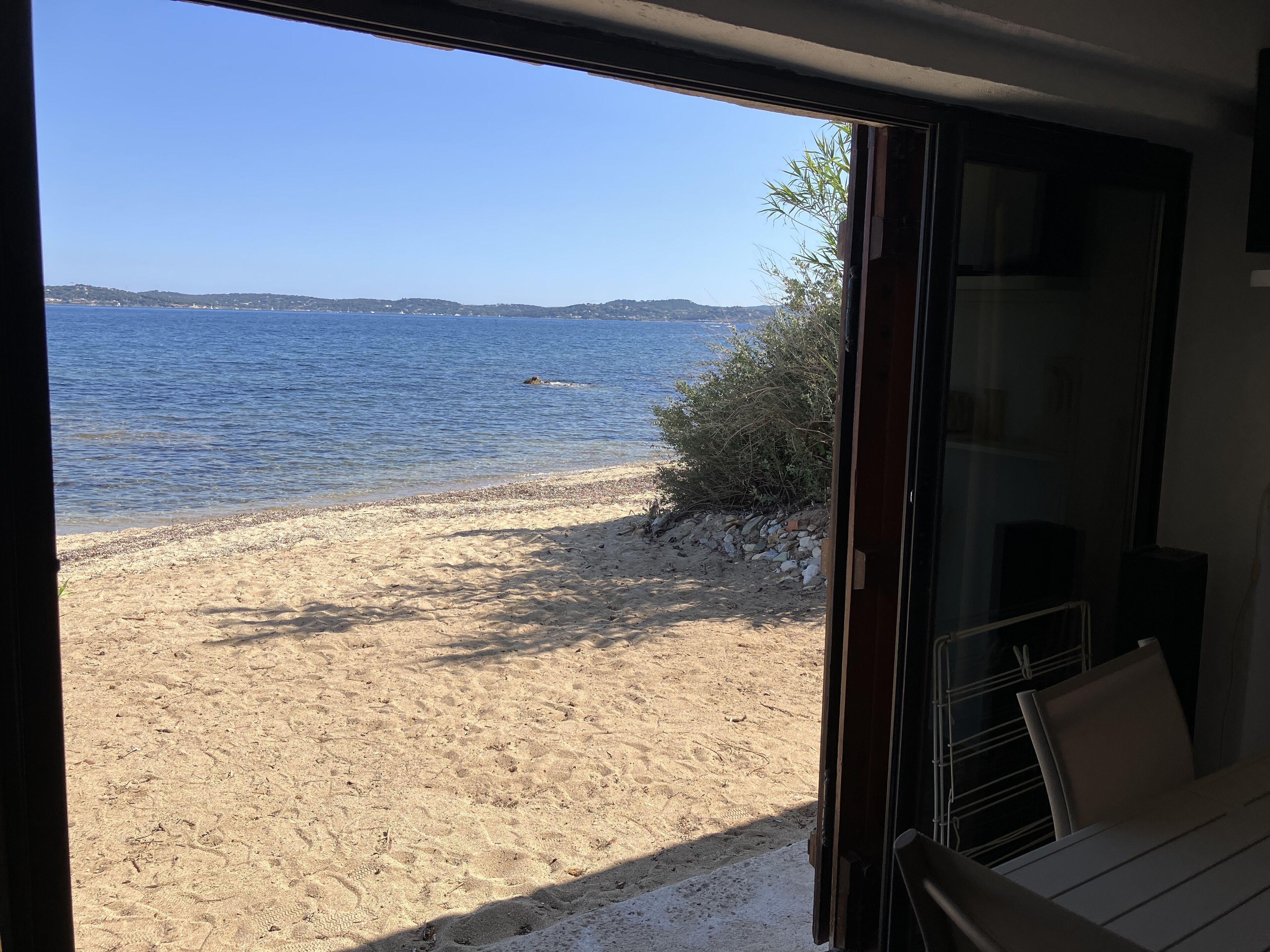 A view from a room with a large window overlooking a sandy beach and calm sea.