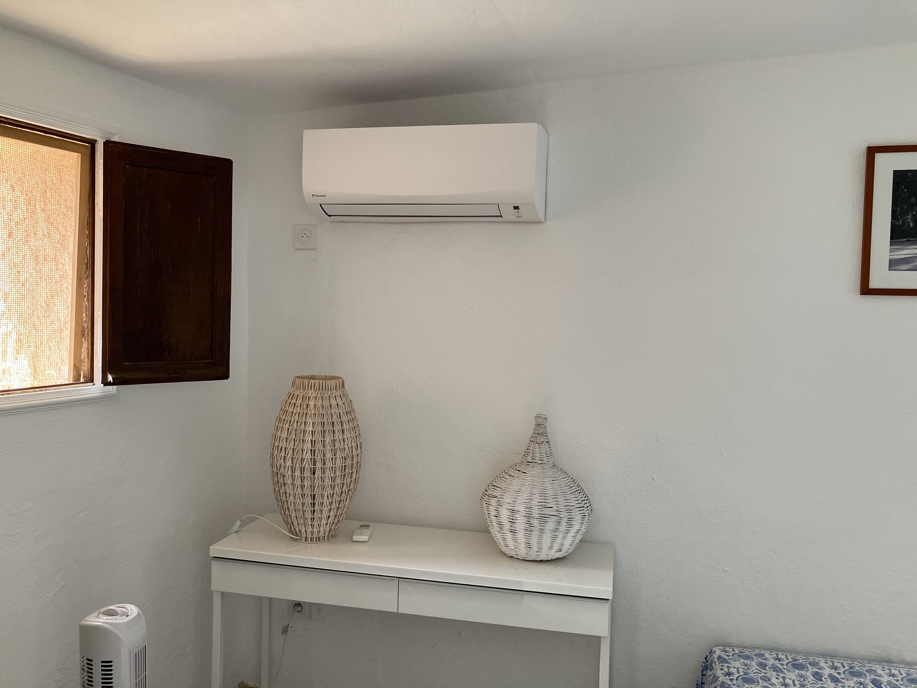 Ein Zimmer mit einer weißen Wand-Klimaanlage, einem weißen Konsolentisch mit zwei dekorativen Vasen und einem Fenster mit einem Holzklappladen.