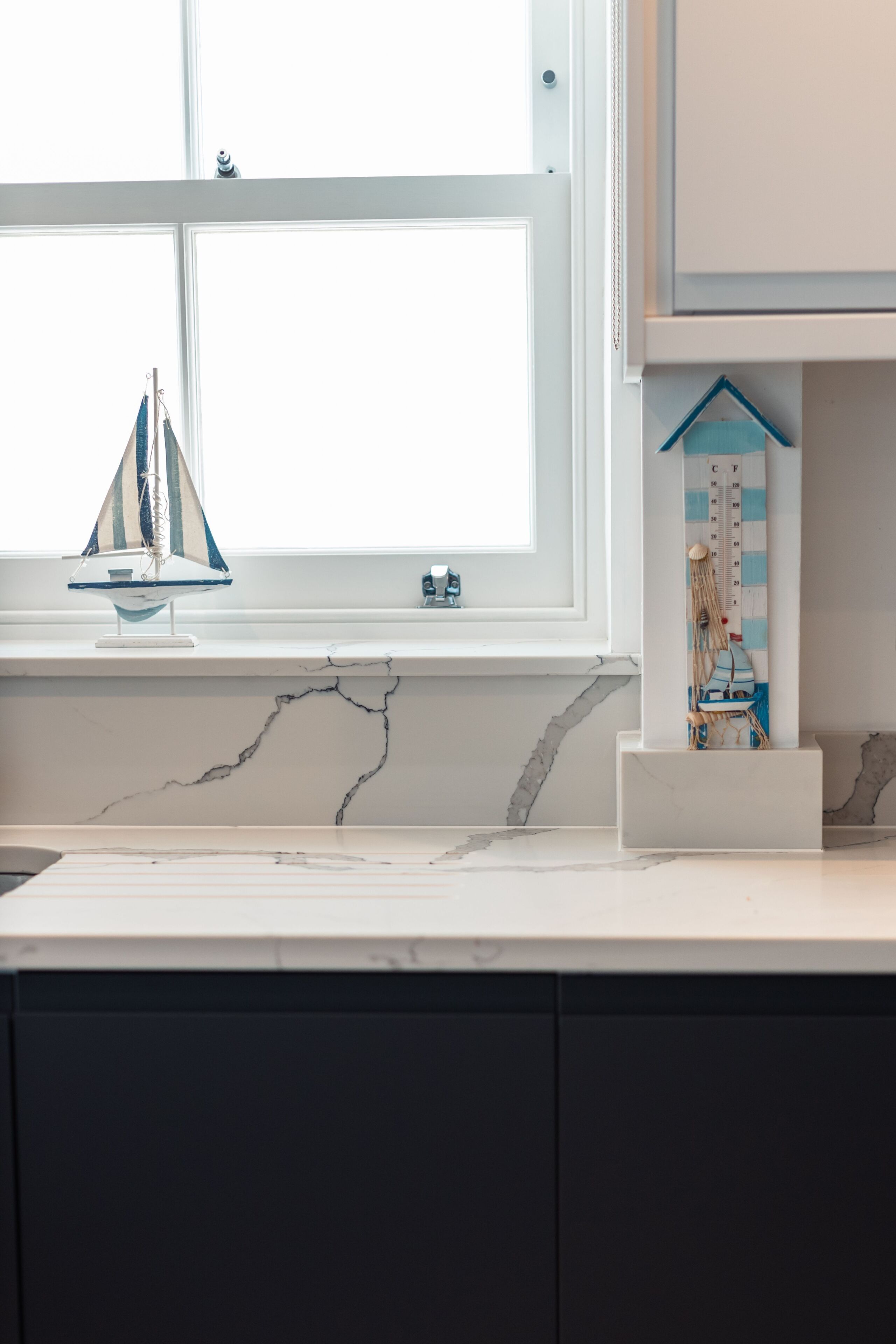 Una cocina con encimera de mármol, una ventana decorada con un velero y un pequeño modelo de casa azul en la estantería.