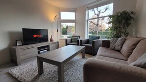 A modern living room with a flat-screen TV displaying Netflix, a sofa, a coffee table, and a large window with a view of a tree and a house.