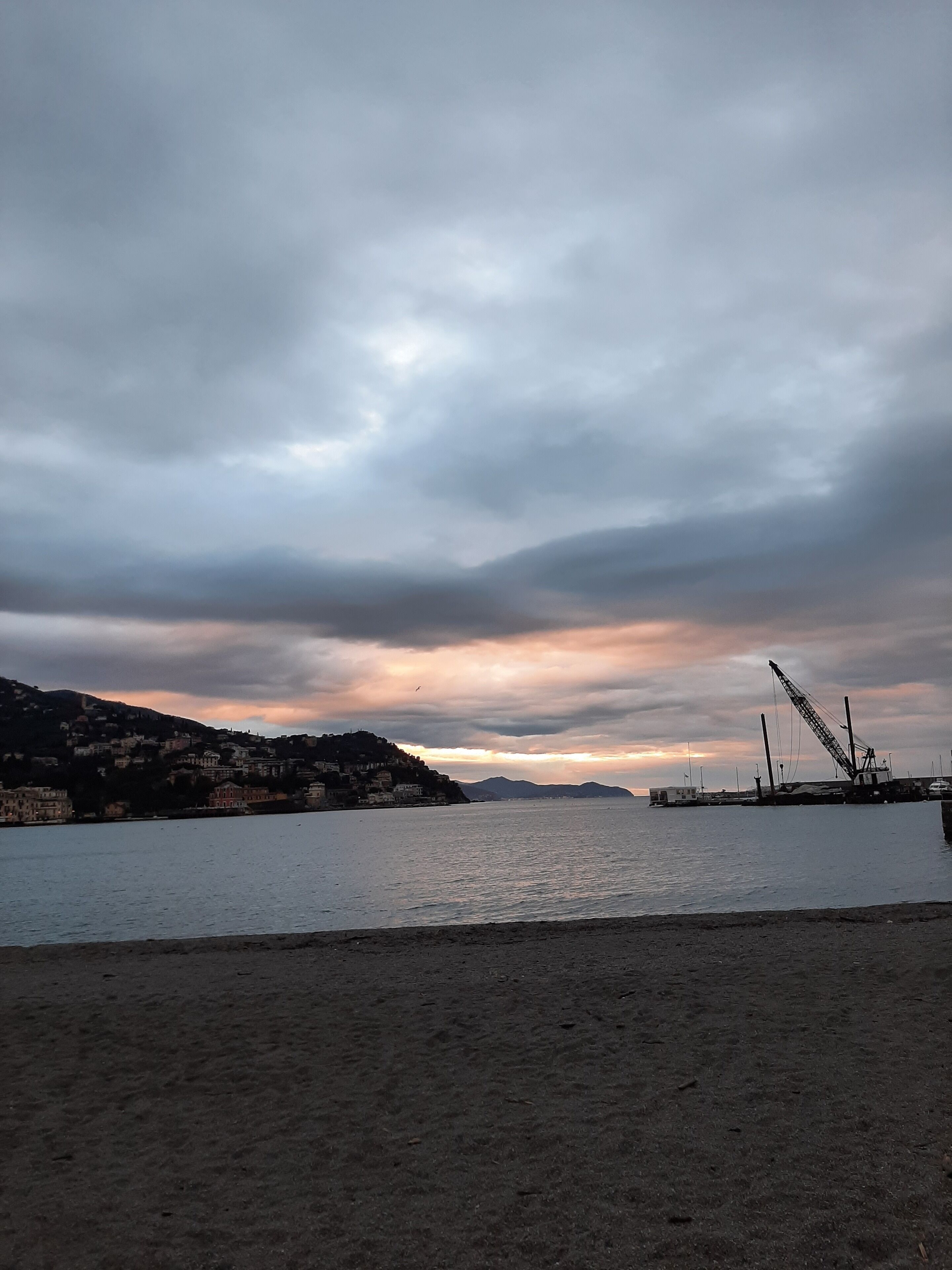 A coastal scene with a calm sea, a sandy beach, and a distant view of a hilly town with buildings.