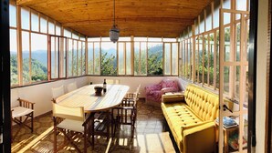 A sunlit room with a wooden ceiling, large windows, a round table, chairs, a yellow sofa, and a hanging light fixture.
