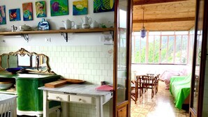 A room with a kitchen area featuring a white tiled backsplash, a wooden countertop, and a green chair. There is a bed with a green blanket and a window with a view of greenery outside.