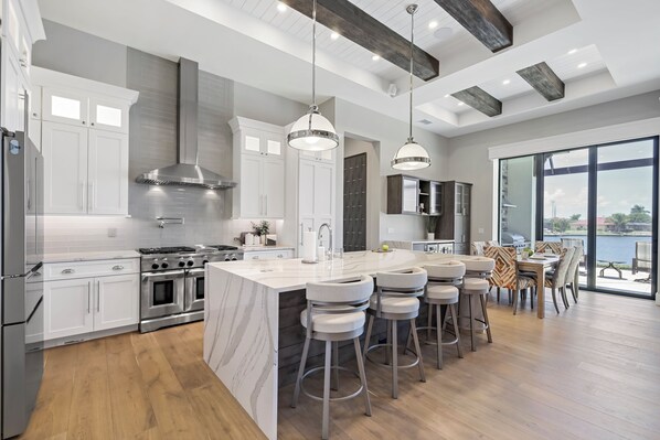 A modern kitchen with stainless steel appliances, a large island with bar stools, and a view of the outdoors through large windows.