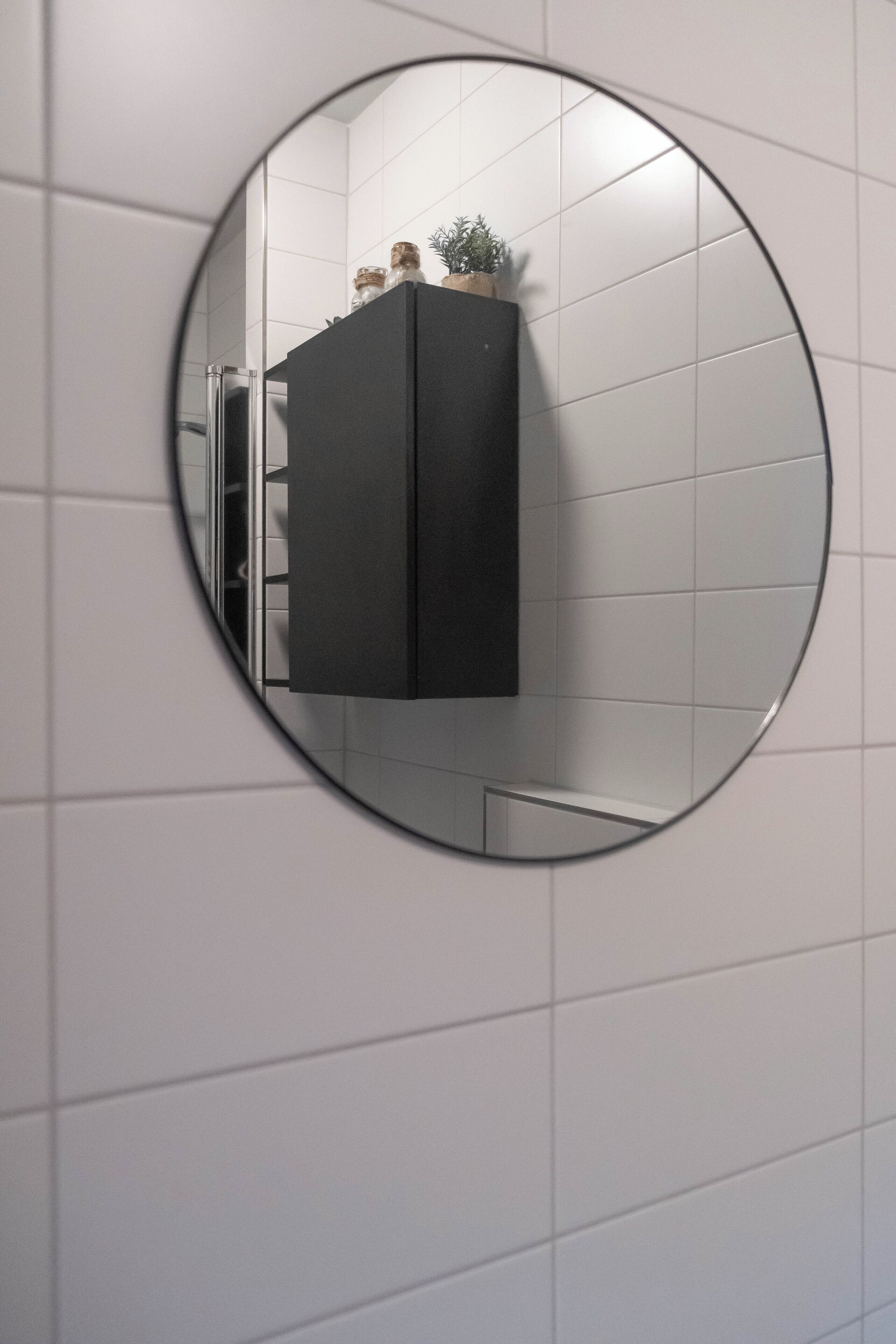 Een ronde spiegel die een zwart kastje weerspiegelt voor een betegelde muur.
