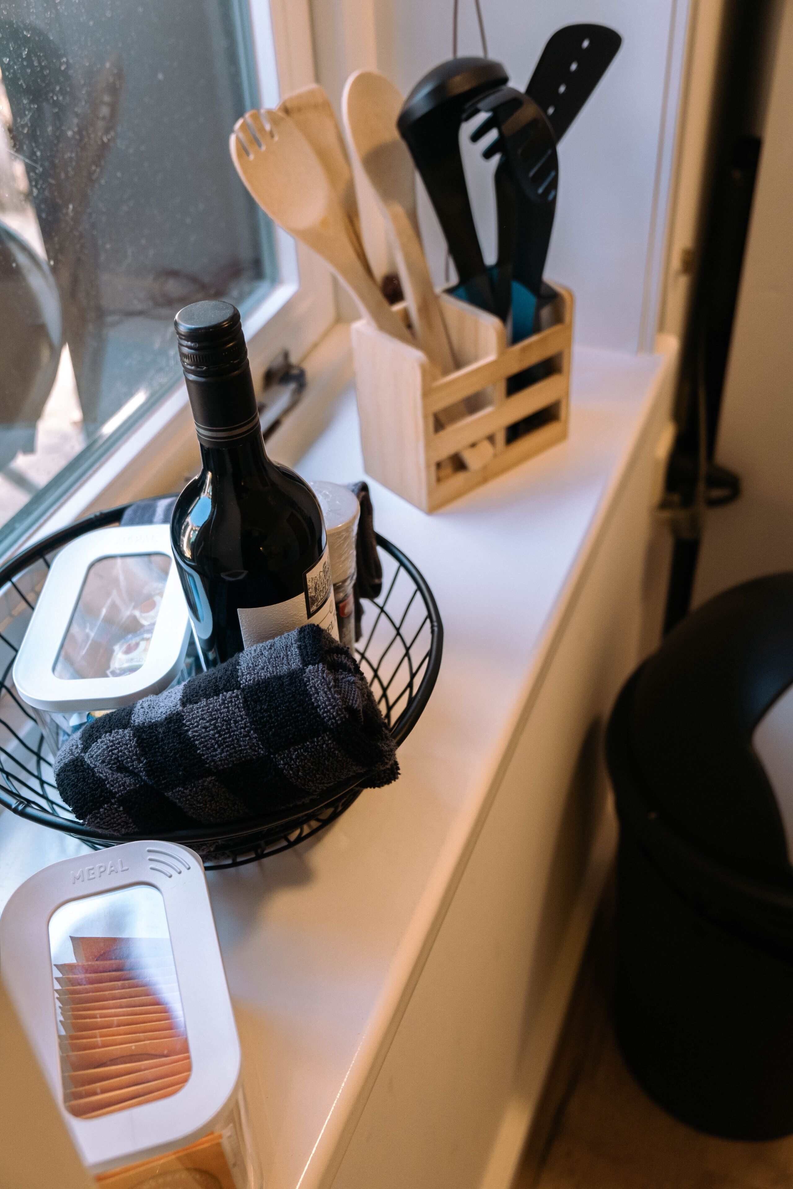 A kitchen counter with a bottle of wine, a basket with a cloth, and a wooden utensil holder.