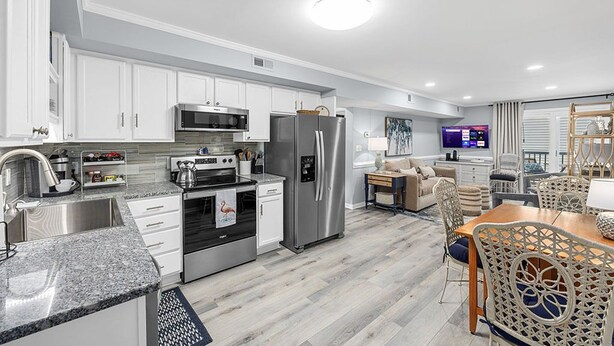 A modern kitchen with stainless steel appliances, white cabinetry, and a dining area with wicker chairs. - Cozy, comfortable and freshly renovated oceanfront condo...you're gonna love it! (North Myrtle Beach)