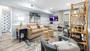 A modern living room with a beige sofa, a coffee table, and a TV. There is a wooden side table with a lamp and a decorative item. The room has a large window with curtains and a painting on the wall. - Cozy, comfortable and freshly renovated oceanfront condo...you're gonna love it! (North Myrtle Beach)