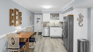 A modern kitchen with white cabinets, a stainless steel refrigerator, and a dining area with a wooden table and wicker chairs. - Cozy, comfortable and freshly renovated oceanfront condo...you're gonna love it! (North Myrtle Beach)