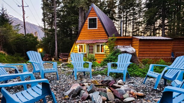 A cabin with a sloped roof surrounded by a gravel area with blue Adirondack chairs. - Rustic Cabin & A-Frame w/ Firepit (Juneau)