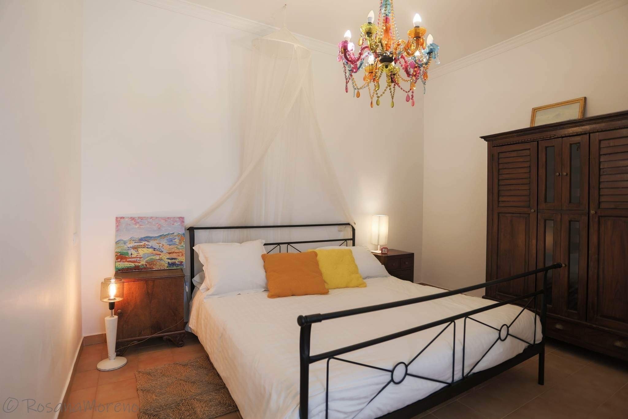 A bedroom with a bed, a chandelier, a nightstand, a cabinet, and a painting on the wall.