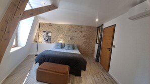 A bedroom with a sloped ceiling, a stone wall, a bed with a patterned bedspread, and a brown ottoman. - Appartement avec terrasse & parking Sarlat Centre (Sarlat-la-Canéda)