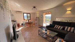 Ein Wohnzimmer mit Sofa, Couchtisch und einer Essecke mit Stühlen und Tisch. Durch ein Fenster ist ein Blick ins Freie möglich.