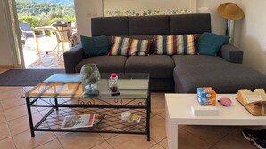 Ein Wohnzimmer mit einer Couch, einem Glastisch, einem weißen Beistelltisch und Blick ins Freie.