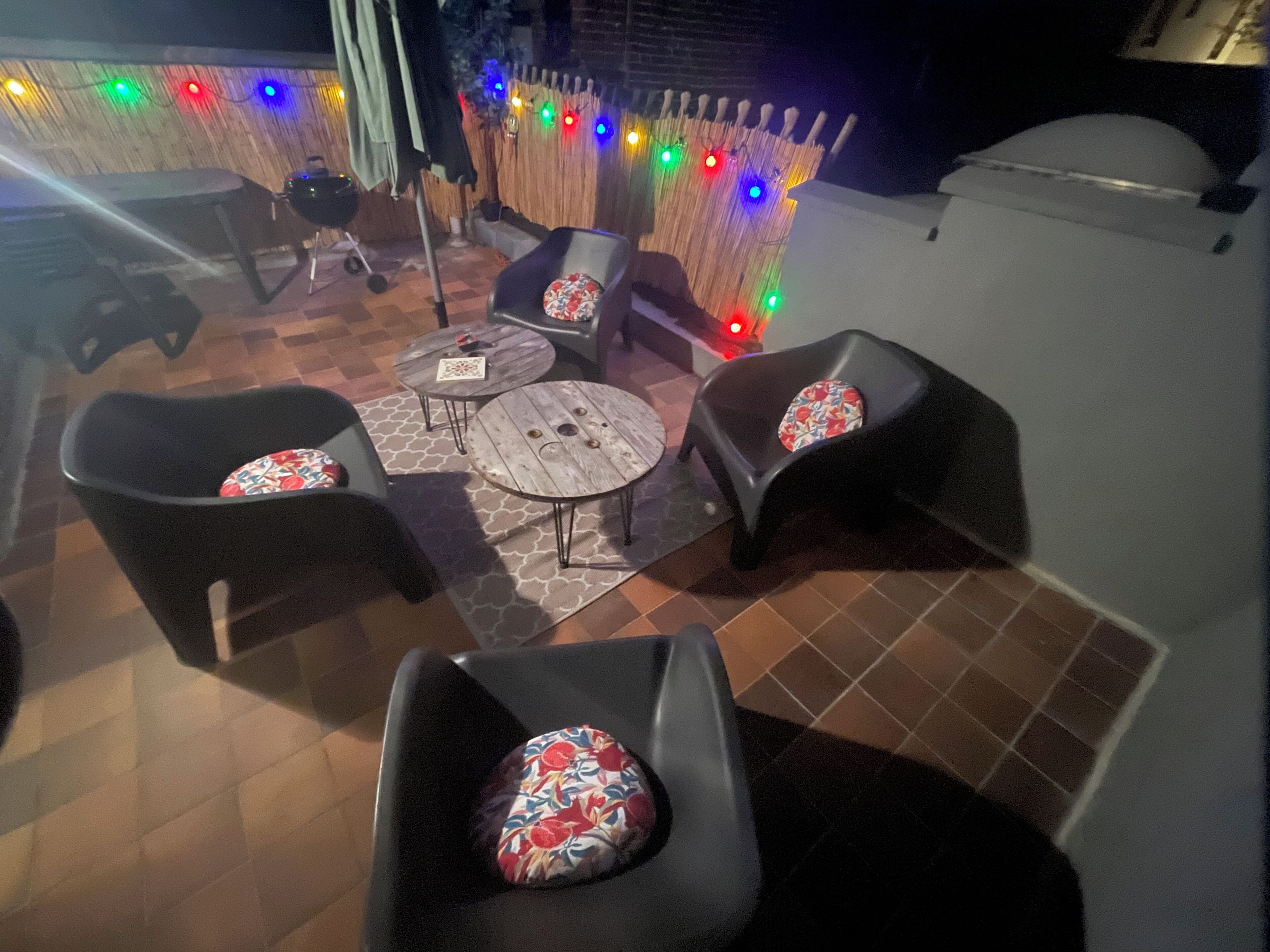 Uma área de pátio externa com quatro cadeiras pretas, duas mesas redondas e luzes de Natal coloridas.