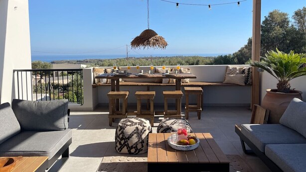 A covered outdoor seating area with a table set for two, a hanging light fixture, and a view of the sea. - Modern Villa with Terrace View, Private Pool & BBQ (Rethymno)