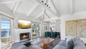 A spacious living room with a fireplace, grey sofas, and a large window overlooking the ocean.