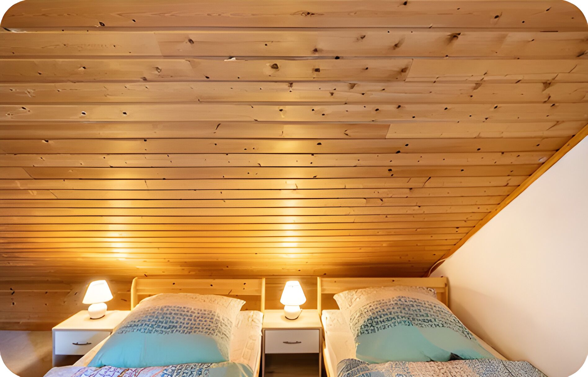 A room with wooden slatted ceiling, two bedside tables with lamps, and two beds with patterned bedding.